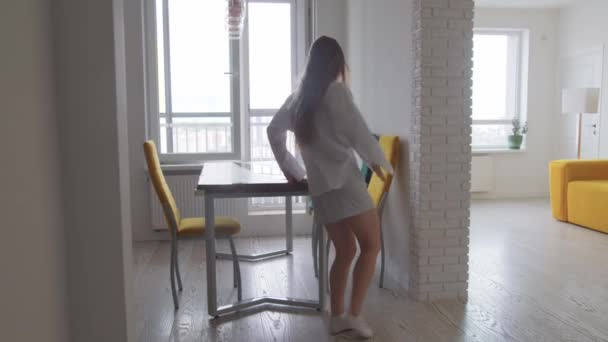 Молода збуджена жінка з довгим волоссям у світлому одязі танцює у своїй світлій квартирі-студії сидить за столом на кухні. Середній постріл
 - Кадри, відео