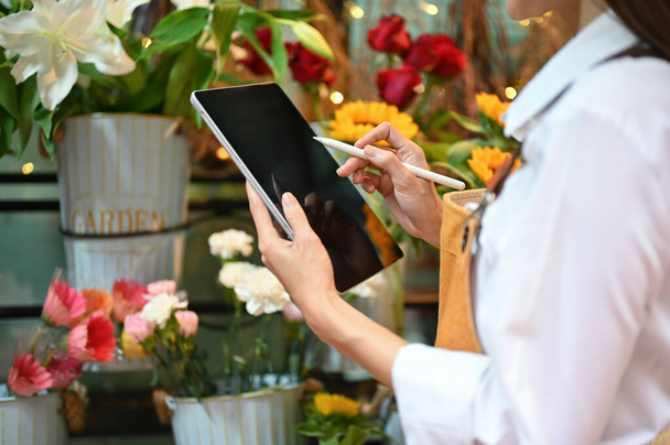 Pretty young Asian female florist or flower shop entrepreneur working in her shop, using digital tablet to check an online order. cropped and close-up image - Fotó, kép