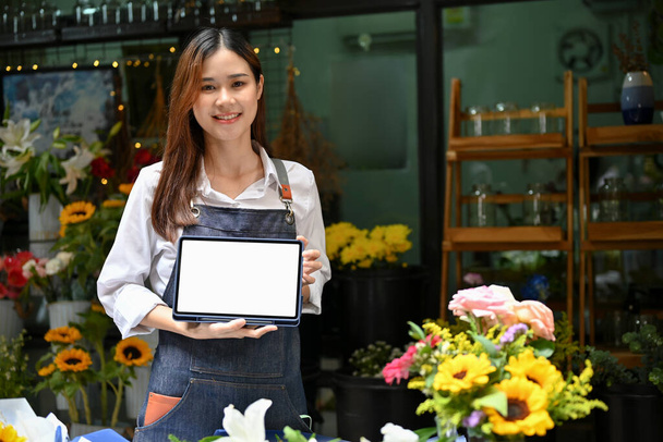Attrayant jeune asiatique fleuriste tenant et montrant une tablette numérique tactile écran blanc maquette, debout devant sa boutique de fleurs. - Photo, image