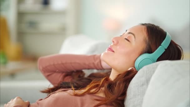 Relajarse, auriculares y mujer feliz escuchando música, radio o podcast mientras descansa en el sofá. Felicidad, sonrisa y chica tranquila de Singapur streaming zen audio en línea en el sofá en la sala de estar en casa - Imágenes, Vídeo