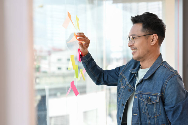 Розумний дорослий азіатський бізнесмен або працівник маркетингу дивиться на записки або липкі банкноти на скляній стіні в своєму офісі.. - Фото, зображення