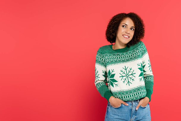 dreamy and smiling african american woman in winter sweater posing with hands in pockets of jeans isolated on red - Foto, Imagen