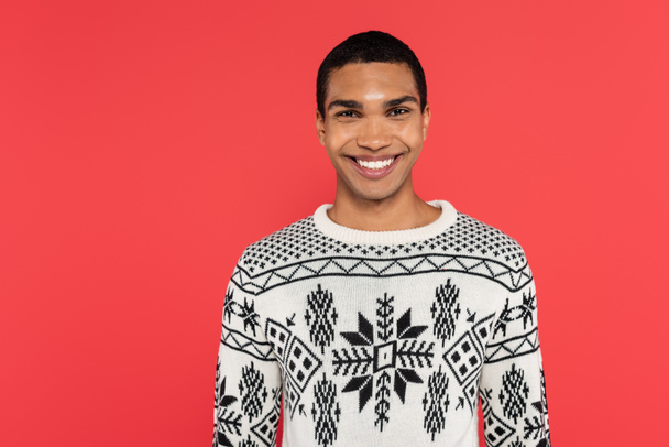 front view of happy african american man in knitted sweater with winter ornament isolated on red - Photo, Image