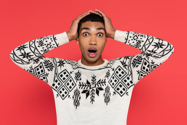 shocked african american man in warm sweater touching head and looking at camera isolated on red - 写真・画像