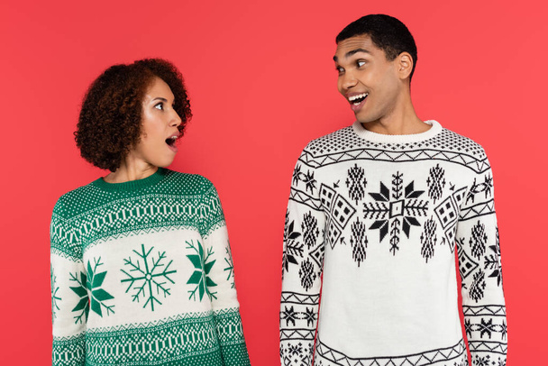 astonished african american couple in warm sweaters looking at each other isolated on red - Foto, Imagen