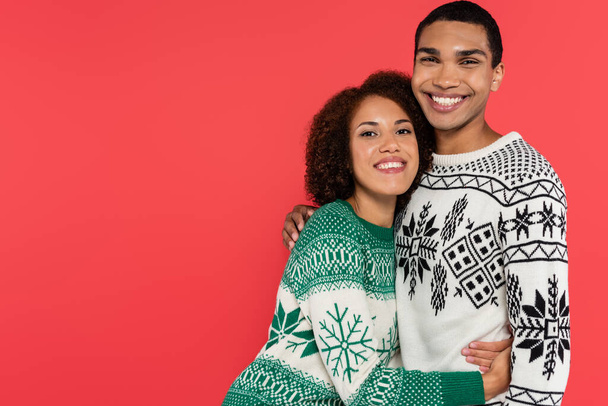 jeune couple afro-américain en chandails avec motif d'hiver embrassant et souriant à la caméra isolée sur rouge - Photo, image