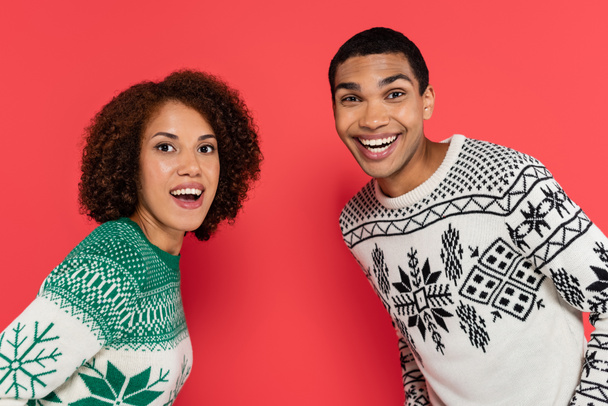 excited african american couple in winter sweaters with ornament looking at camera isolated on red - Фото, изображение