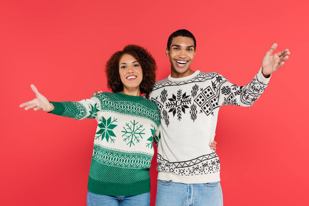 happy african american couple in warm sweaters with winter ornament showing greeting gesture isolated on red - Φωτογραφία, εικόνα