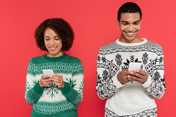 usmívající se africký americký pár ve svetrech se zimním vzorem chatování na smartphonech izolovaných na červené - Fotografie, Obrázek