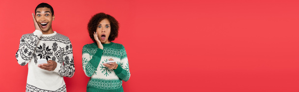 shocked african american woman in winter sweaters holding cellphones and looking at camera isolated on red, banner - Valokuva, kuva