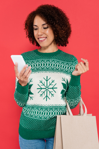 donna afroamericana sorridente in maglione con motivo invernale in piedi con shopping bag e smartphone isolati su rosso - Foto, immagini