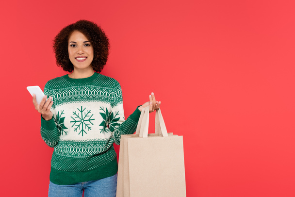 cheerful african american woman in warm sweater holding shopping bags and smartphone isolated on red - Valokuva, kuva