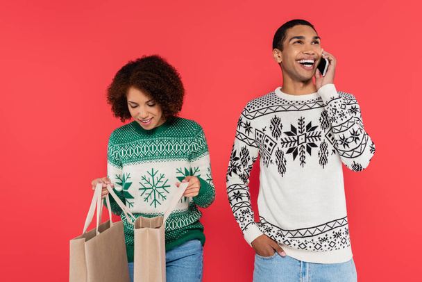 african american woman looking into shopping bags near cheerful man talking on cellphone isolated on red - Foto, Imagem