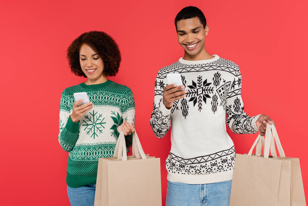 feliz pareja afroamericana con teléfonos móviles y bolsas de compras aisladas en rojo - Foto, imagen