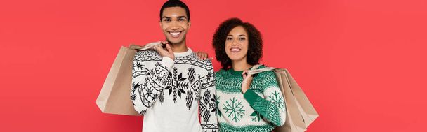 complacida pareja afroamericana en suéteres con patrón de invierno sosteniendo bolsas aisladas en rojo, pancarta - Foto, imagen