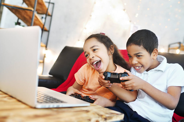two African American children - a boy and a girl holding a gamepad in their hands, play computer games on a laptop and laugh while sitting on the couch. - Foto, imagen