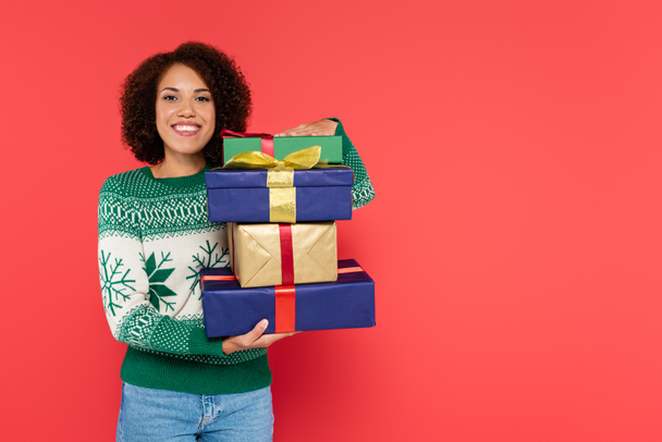 pleased african american woman in winter sweater holding stack of gift boxes and looking at camera isolated on red - Фото, изображение