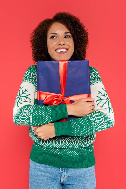 heureuse femme afro-américaine en pull d'hiver étreignant boîte cadeau bleue avec ruban lumineux isolé sur rouge - Photo, image