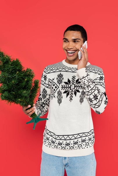 radostný africký Američan ve svetru se zimní ozdobou drží malé vánoční stromeček, zatímco mluví na mobilní telefon izolované na červené - Fotografie, Obrázek