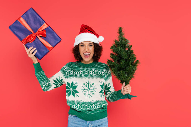 mulher americana africana alegre em chapéu de santa e suéter com padrão de inverno segurando pequena árvore de natal e caixa de presente isolado no vermelho - Foto, Imagem
