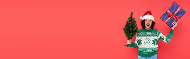donna afroamericana eccitata in maglione e cappello di Babbo Natale tenendo presente e albero di Natale isolato su rosso, banner - Foto, immagini
