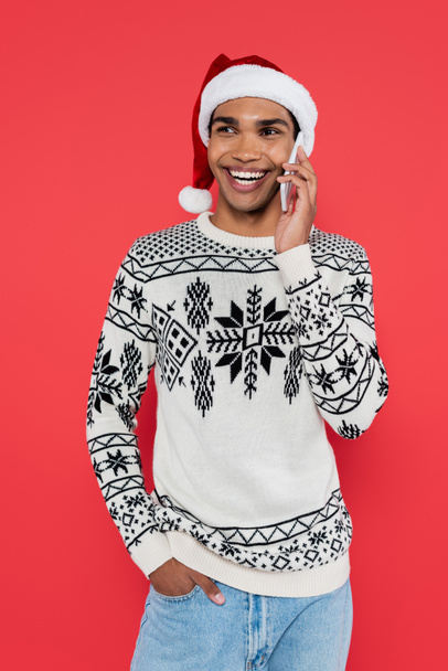 smiling african american man in santa hat standing with hand in pocket while talking on smartphone isolated on red - Photo, Image