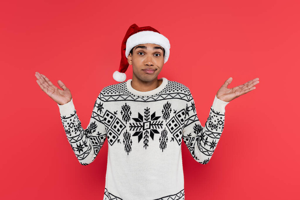 discouraged african american man in santa hat and white sweater with winter pattern showing shrug gesture isolated on red - Foto, imagen