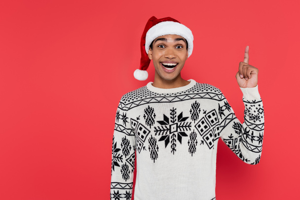 cheerful african american man in santa hat and winter sweater showing idea gesture isolated on red - Foto, imagen
