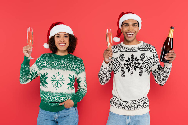 joyful african american couple in santa hats holding champagne glasses and looking at camera isolated on red - Фото, зображення