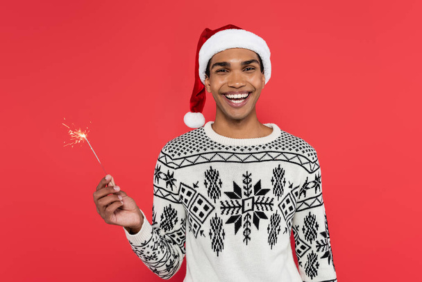 joyful african american man in santa hat and winter sweater holding sparkler and looking at camera isolated on red - Φωτογραφία, εικόνα