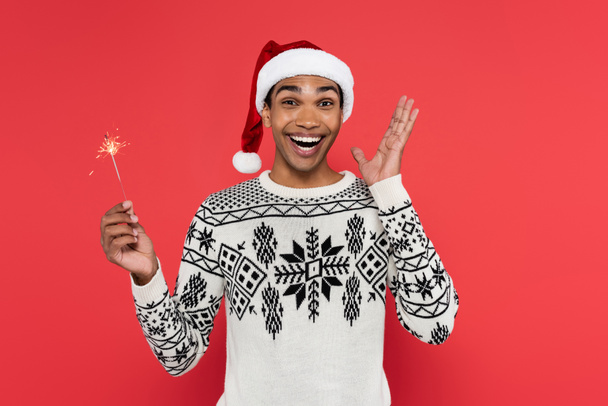 excited african american man in santa hat holding sparkler and showing wow gesture isolated on red - Foto, imagen