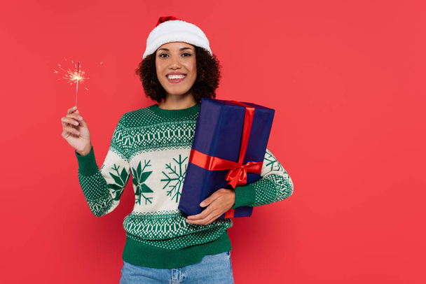 joyful african american woman in santa hat and winter sweater holding sparkler and blue gift box isolated on red - Valokuva, kuva