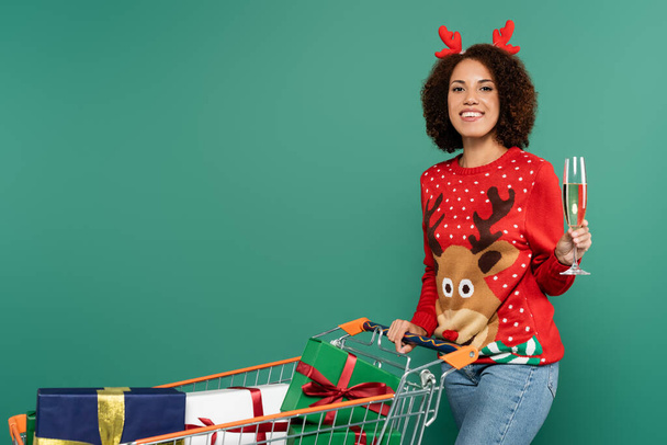 mulher afro-americana em roupa de Natal segurando taça de champanhe perto do carrinho de compras com presentes isolados em verde - Foto, Imagem