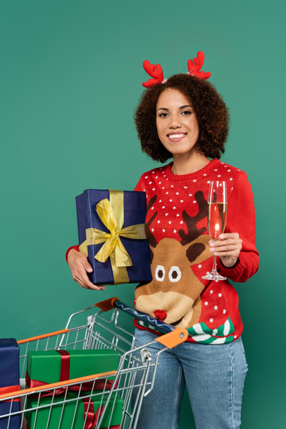 happy african american woman in festive outfit holding champagne glass and present near shopping cart isolated on green - Photo, Image