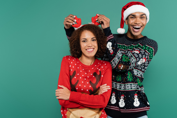 excited african american man in santa hat touching reindeer horns on headband of girlfriend isolated on green - Photo, Image