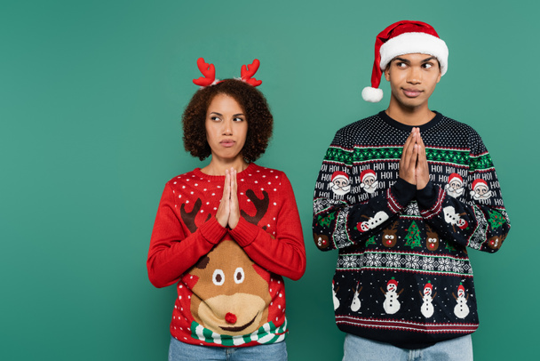 coppia afro-americana tesa in abito natalizio in piedi con le mani in preghiera e guardando lontano isolato sul verde - Foto, immagini