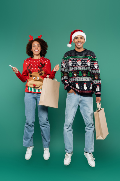 heureux couple afro-américain en tenue de Noël lévitant avec téléphone portable et sacs à provisions sur fond vert - Photo, image