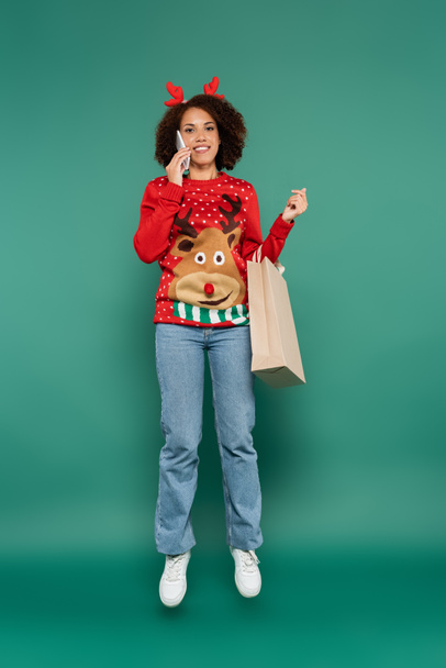 african american woman in christmas outfit talking on smartphone while levitating with shopping bag on green background - Foto, immagini