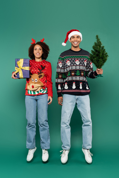 happy african american couple in festive sweaters levitating with gift box and small christmas tree on green background - 写真・画像