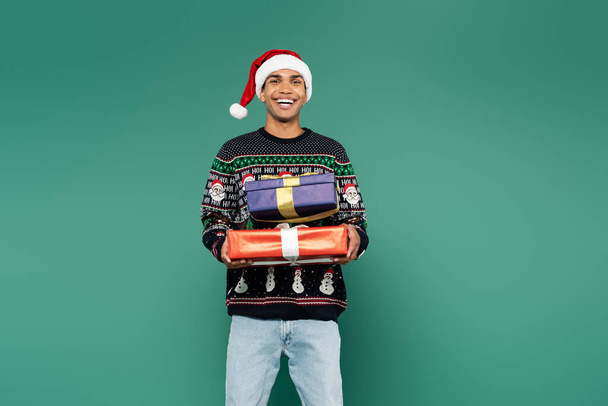excited african american man in santa hat and winter sweater holding christmas presents isolated on green - Photo, Image