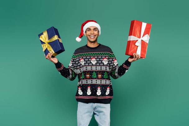 pleased african american man in santa hat and sweater with festive pattern holding gift boxes isolated on green - Fotó, kép