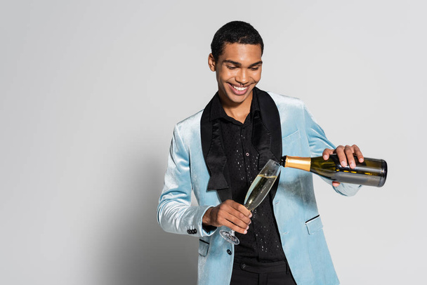 joyful african american man in elegant silk jacket pouring champagne on grey background - Fotó, kép