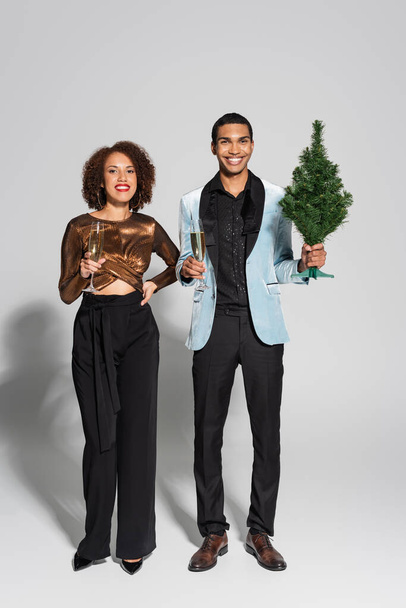 volledige lengte van elegant en stijlvol Afrikaans Amerikaans paar met champagne en kleine kerstboom op grijze achtergrond - Foto, afbeelding