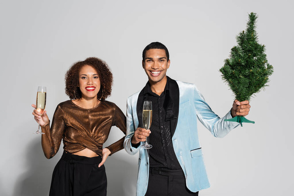 Стильна афро-американська пара з шампанськими окулярами і маленькими кришталевими деревами на сірому тлі.   - Фото, зображення