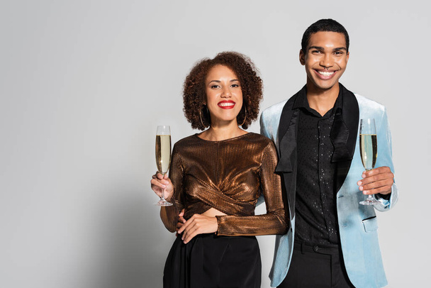 african american woman in shiny blouse and man in silk blazer holding champagne glasses on grey background - Photo, Image