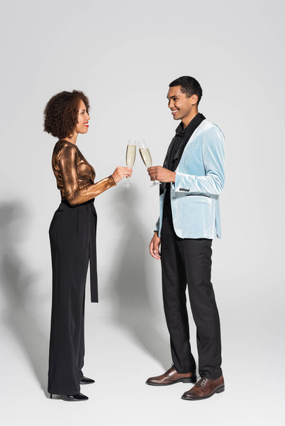 boczny widok eleganckiej afrykańskiej pary trzymającej kieliszki do szampana i uśmiechającej się do siebie na szarym tle - Zdjęcie, obraz