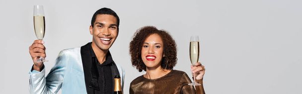 стильная и элегантная африканская пара с бокалами для шампанского, улыбающаяся в камеру, изолированную на сером, баннере - Фото, изображение