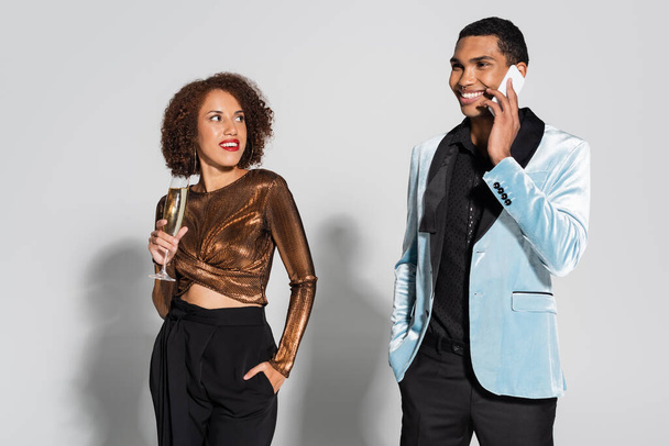 smiling african american man in silk blazer talking on smartphone near elegant woman with champagne glass on grey background - Foto, imagen