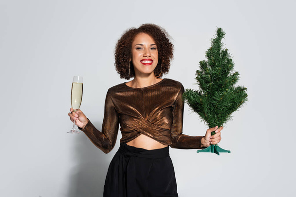 stylish african american woman with small christmas tree and champagne glass smiling at camera on grey background - Photo, Image