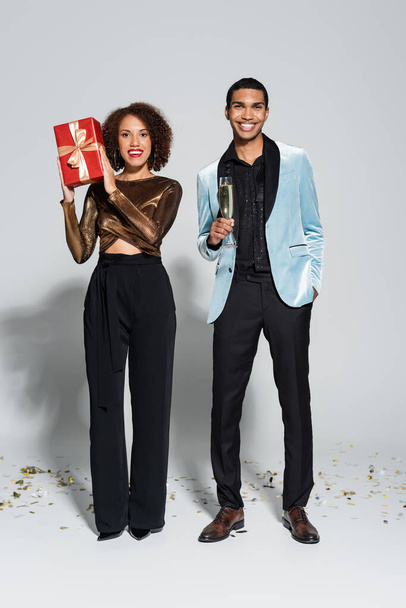 elegant african american man with champagne glass and hand in pocket near girlfriend with christmas present on grey background - Photo, image